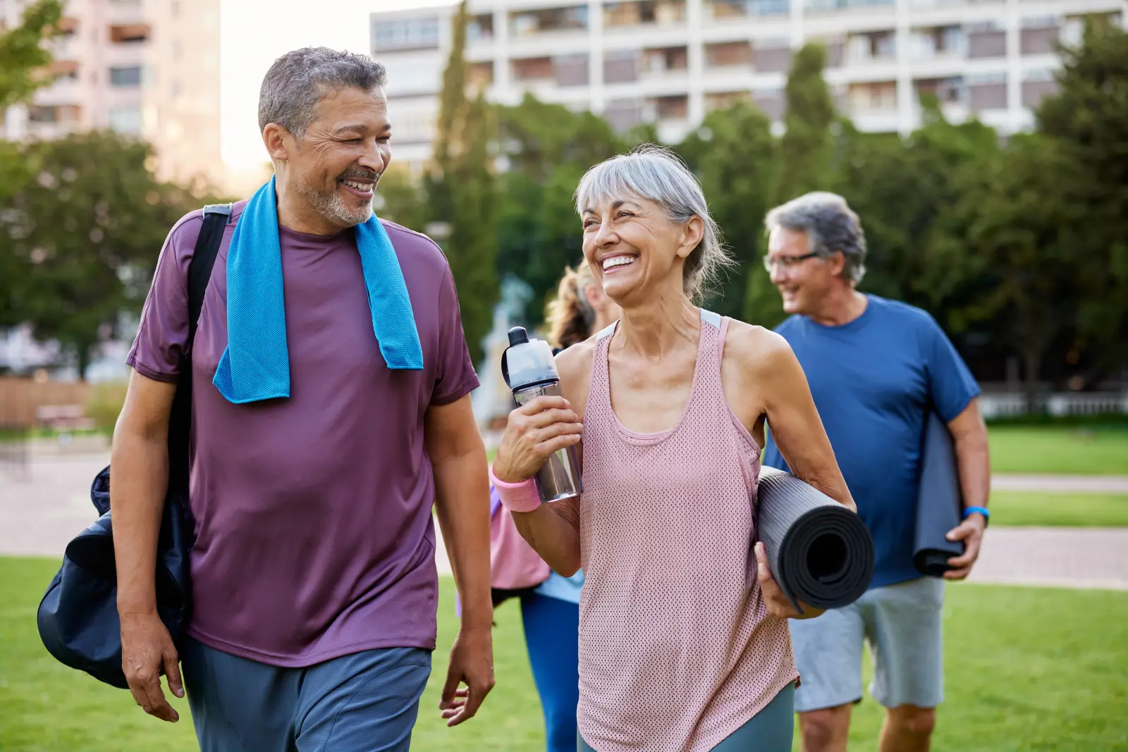 Smiling older adults walk across a park after an exercise class, carrying yoga mats and a water bottle.